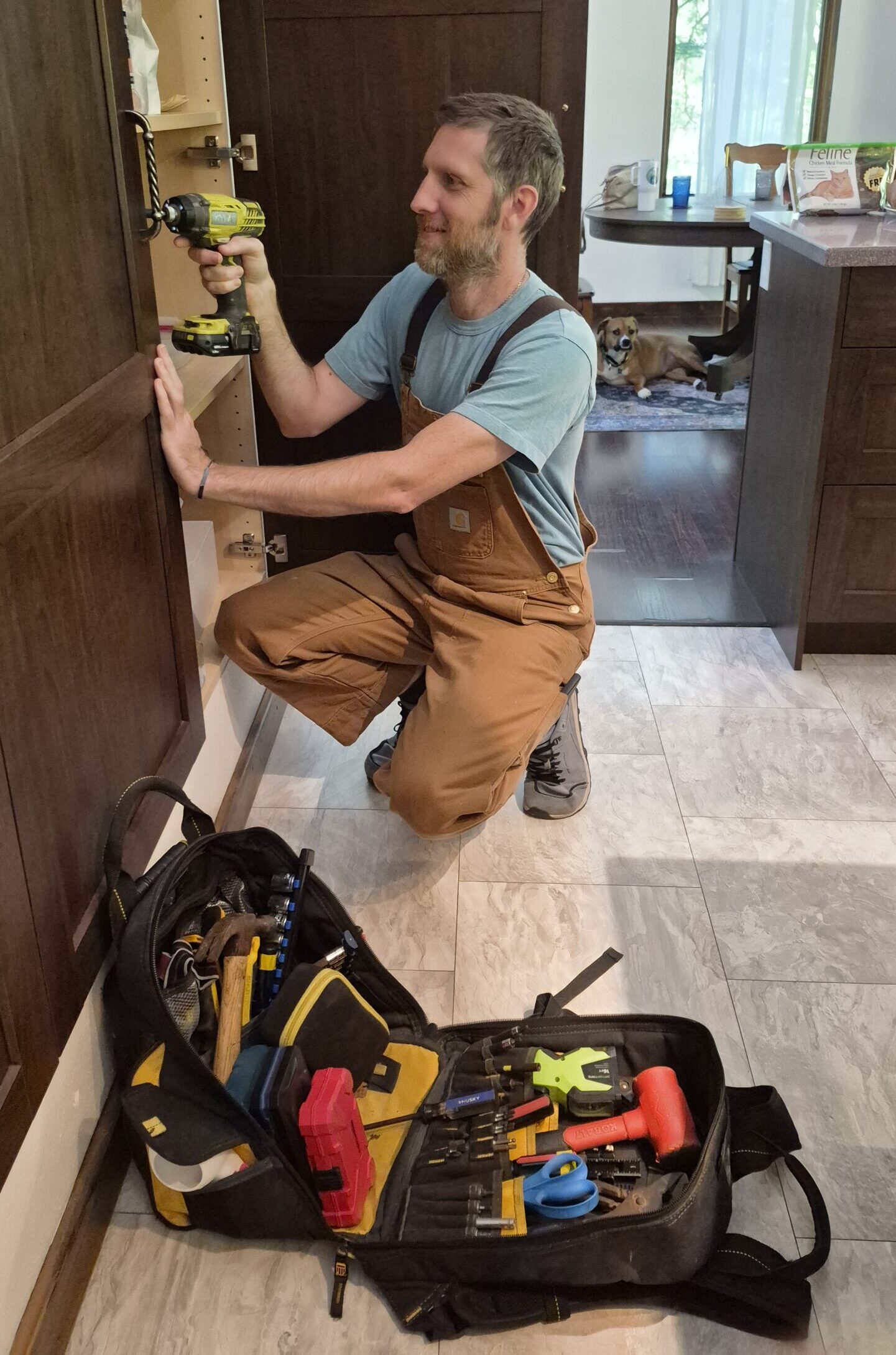 Marlon is crouching to use a power drill on a large cabinet. There is a content dog in the background.